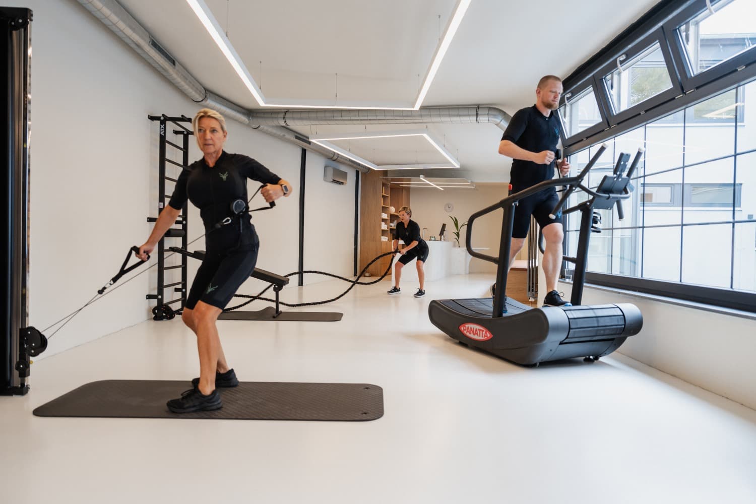 Ein Bild von 3 Personen im EMS-Anzug von Antelope in einem Fitnessstudio. Alle führen unterschiedliche Übungen aus.